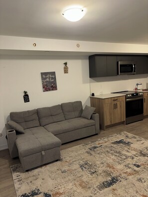 A living room with a grey sectional sofa, a kitchen with dark cabinets and a microwave, and a patterned rug.
