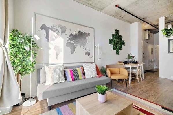 Un salon moderne avec un canapé gris, des coussins colorés, une carte du monde accrochée au mur, un coin repas avec une table et des chaises, et une plante en pot posée sur une table basse.