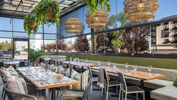 An outdoor dining area with wicker furniture, hanging plants, and large windows offering views of the street. - Eminence Sleek Studio City View (Edmonton)