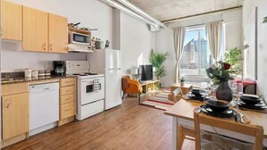 A compact kitchen with wooden cabinets, a microwave, and a stove. There is a dining area with a table set for two and a TV in the background. - Eminence Sleek Studio City View (Edmonton)
