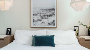 Une chambre à coucher avec un lit, des oreillers blancs, un coussin décoratif de couleur turquoise, un tableau encadré représentant un paysage marin et des lampes suspendues.