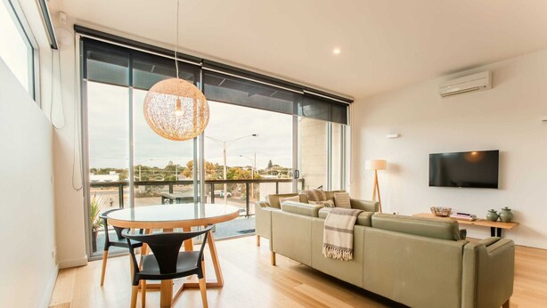 Un salon moderne comprenant un canapé, une table à manger avec des chaises, un luminaire suspendu et un téléviseur fixé au mur.