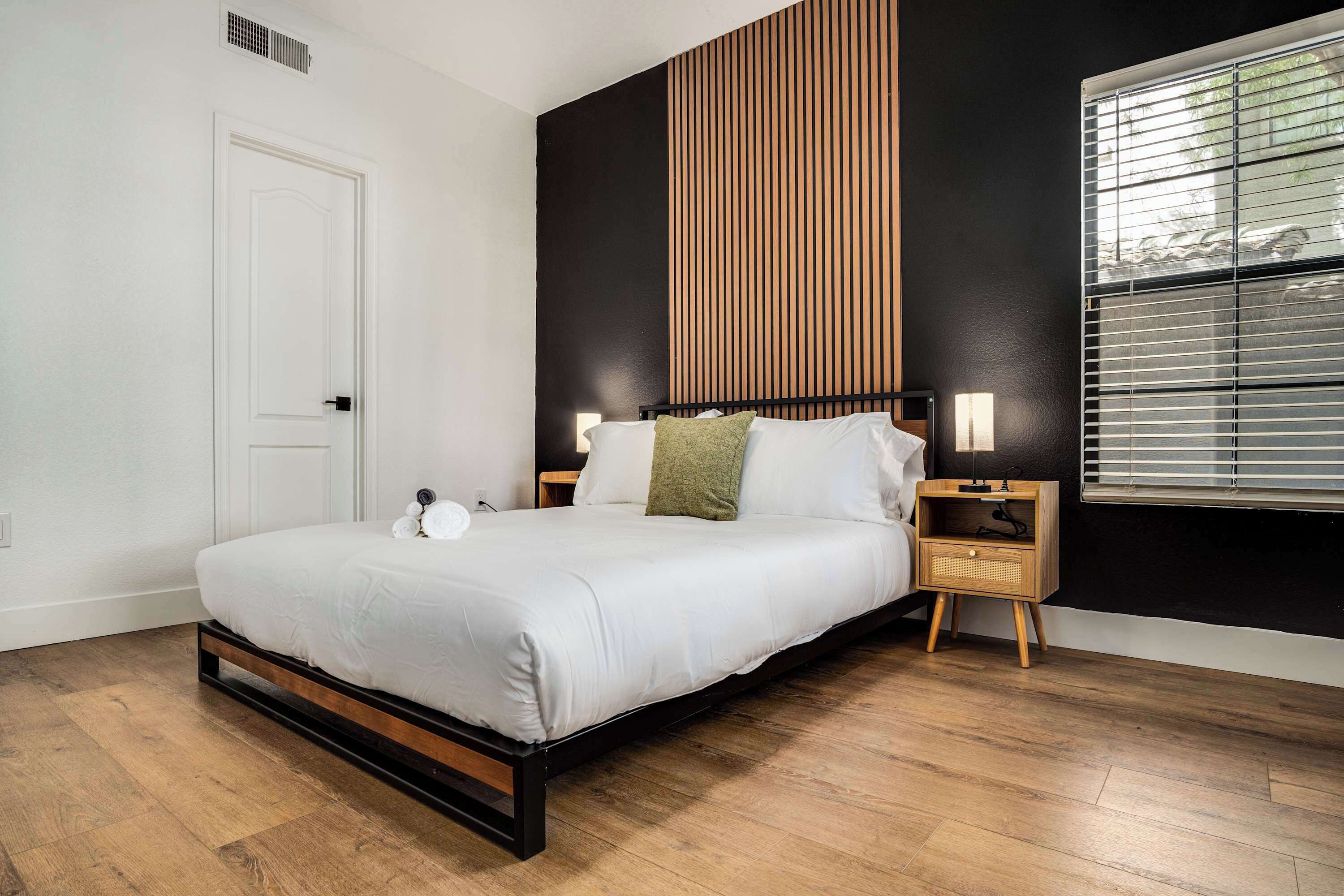 A modern bedroom with a wooden floor, a white bed with a black frame, two bedside tables with lamps, and a window with blinds.
