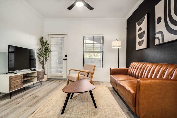 A living room with a flat-screen TV, a wooden entertainment unit, a brown leather sofa, a wooden chair, and a floor lamp. - Boulevard Stays Quiet Modern 1BR Pickleball, Pool, Parking, Gym 07 (Chandler)