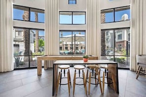 A spacious room with a large table and four stools, large windows with a view of a pool and buildings, and a stone accent wall.
