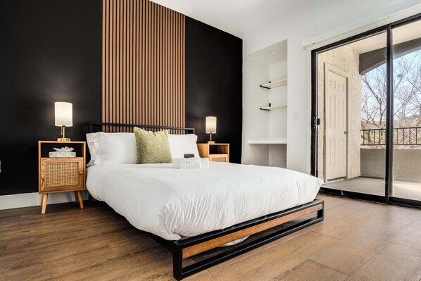A modern bedroom with a large bed, wooden headboard, bedside tables with lamps, and a sliding glass door to a balcony.