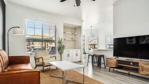 A modern living room with a sofa, coffee table, TV, and kitchen area. - Boulevard Stays 2BR by Stadium Pool, Parking, Gym 10 (Glendale)