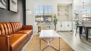 A modern living room with a leather sofa, a white table, and a kitchen area with white cabinets and a window. - Boulevard Stays 2BR by Stadium Pool, Parking, Gym 10 (Glendale)
