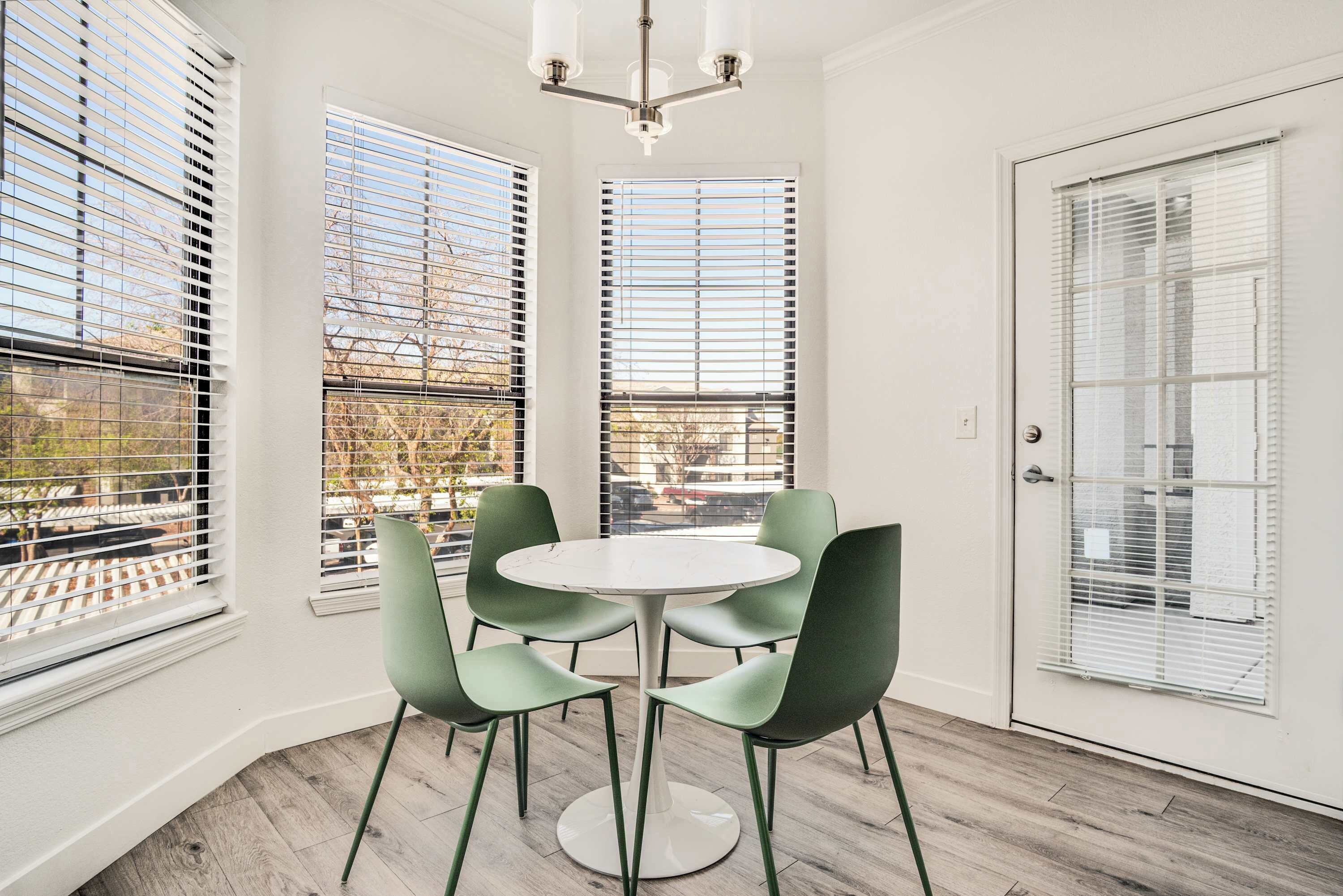 A room with a round table and four chairs, large windows with blinds, a door, and a chandelier.