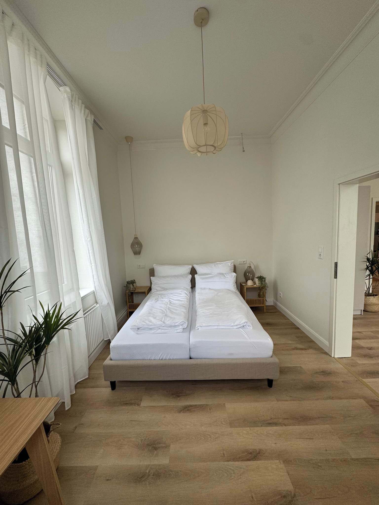 Un dormitorio con una cama grande, cortinas blancas, un lámpara colgante y suelo de madera.
