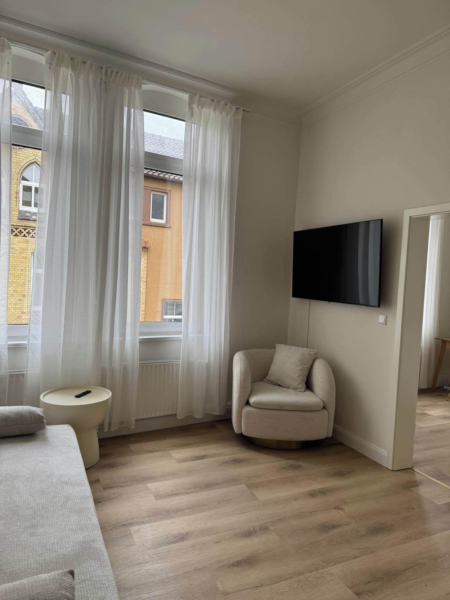 A modern living room with a sofa, a round armchair, a TV mounted on the wall, and a window with sheer curtains.