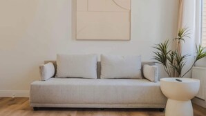 Un salon avec un canapé de couleur claire, un tableau abstrait moderne, une plante en pot et une fenêtre avec des rideaux.