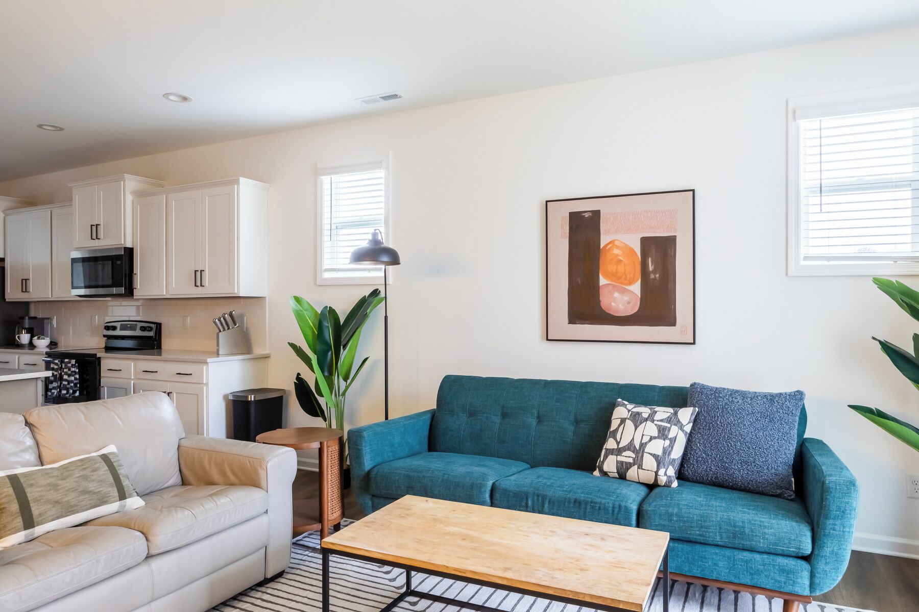 A modern living room with a teal sofa, a white leather couch, and a wooden coffee table. A kitchen with white cabinets and appliances is visible in the background.