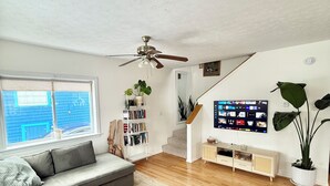 A modern living room with a grey sofa, a ceiling fan, a bookshelf, a TV, and a staircase.