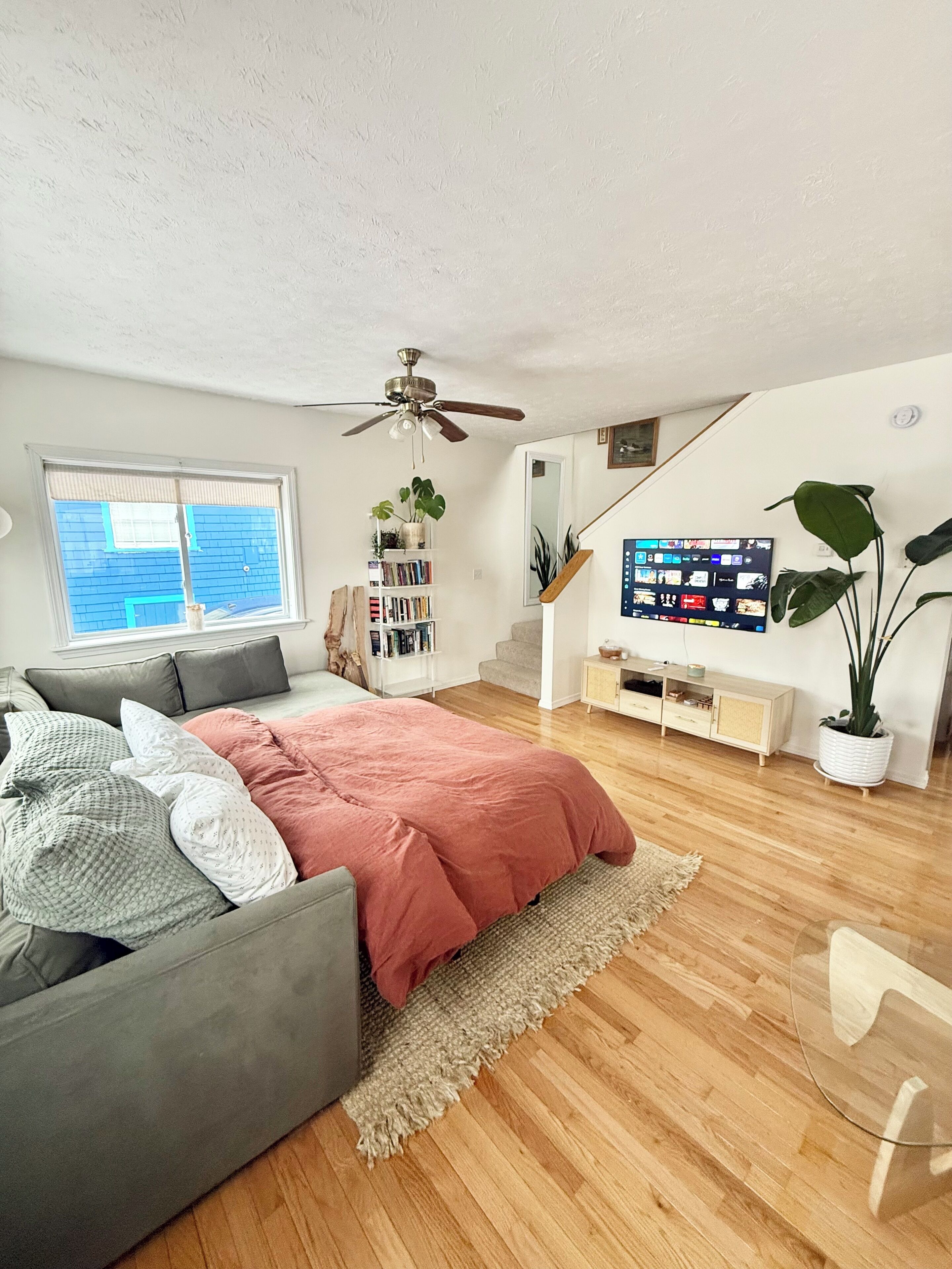 Una camera da letto moderna con un letto, un divano grigio, pavimento in legno, una ventola a soffitto, una libreria, un televisore e un'ampia finestra.