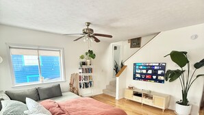 A modern bedroom with a bed, a grey sofa, a wooden floor, a ceiling fan, a bookshelf, a TV, and a large window.