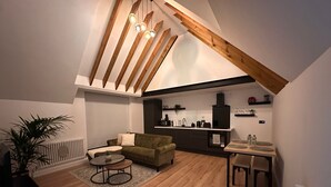 Un loft moderne avec de hauts plafonds, des poutres apparentes, un coin salon confortable avec un canapé vert et une table basse, une cuisine avec des meubles sombres et une petite table à manger.