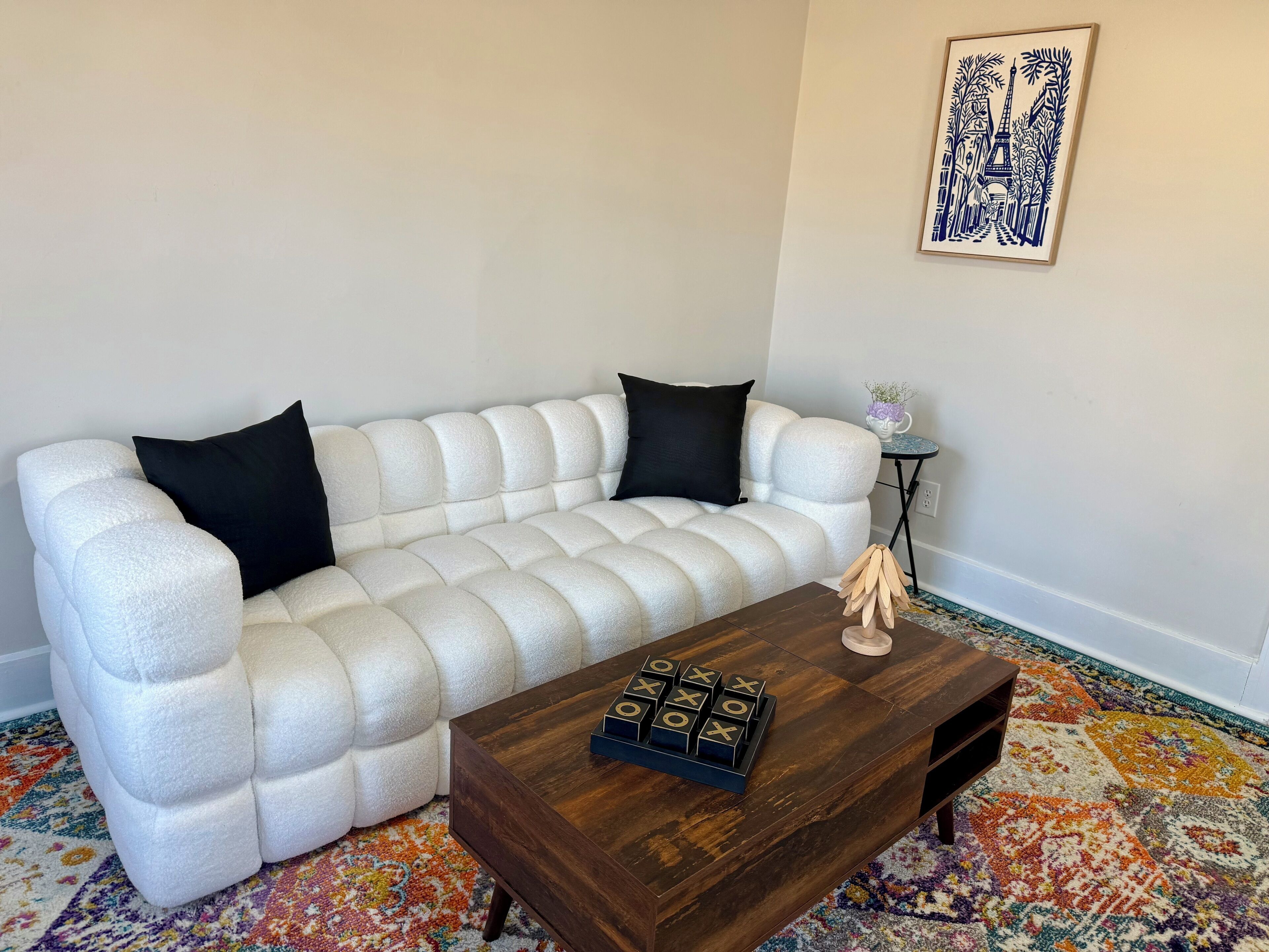 Ein weißes, gepolstertes Sofa mit schwarzen Kissen, ein Couchtisch aus Holz mit einem Gesellschaftsspiel, ein farbenfroher Teppich mit Muster und ein gerahmtes Kunstwerk an der Wand.