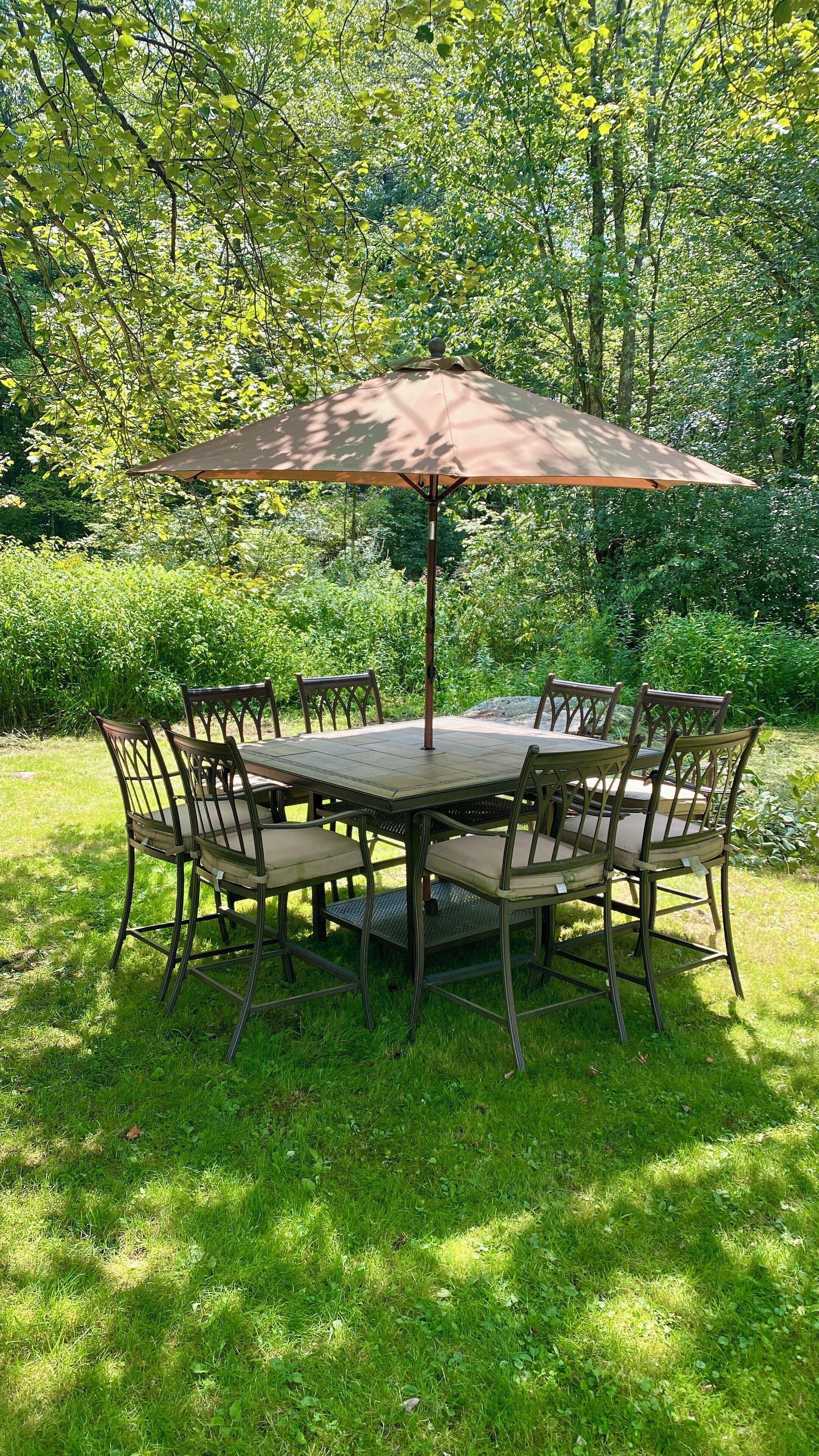 Une table ronde entourée de six chaises, et un grand parasol, le tout installé dans un jardin luxuriant.