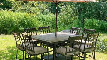 Une table ronde entourée de six chaises, et un grand parasol, le tout installé dans un jardin luxuriant.