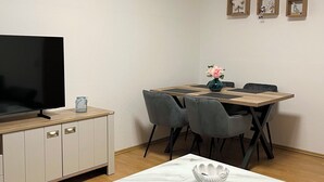 Uma sala de estar moderna com televisão de tela plana, uma mesa de jantar de madeira com cadeiras, uma mesa de centro de mármore e um pequeno vaso decorativo.