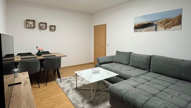 Uma sala de estar moderna com um sofá modular cinza, uma mesa de jantar de madeira com cadeiras, uma pequena mesa de centro branca e um tapete cinza.