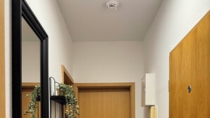Um corredor com porta de madeira, espelho com moldura preta, uma planta pequena, armário branco e uma estante de metal.