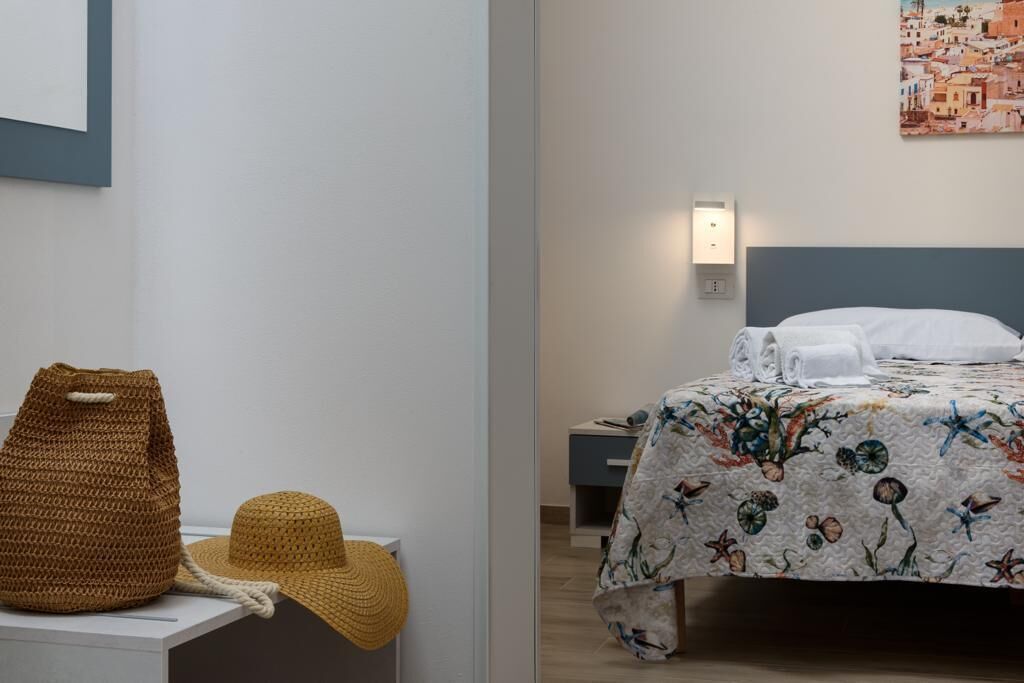 A bedroom with a bed, a nightstand, a wall-mounted light, and a painting of a coastal town.
