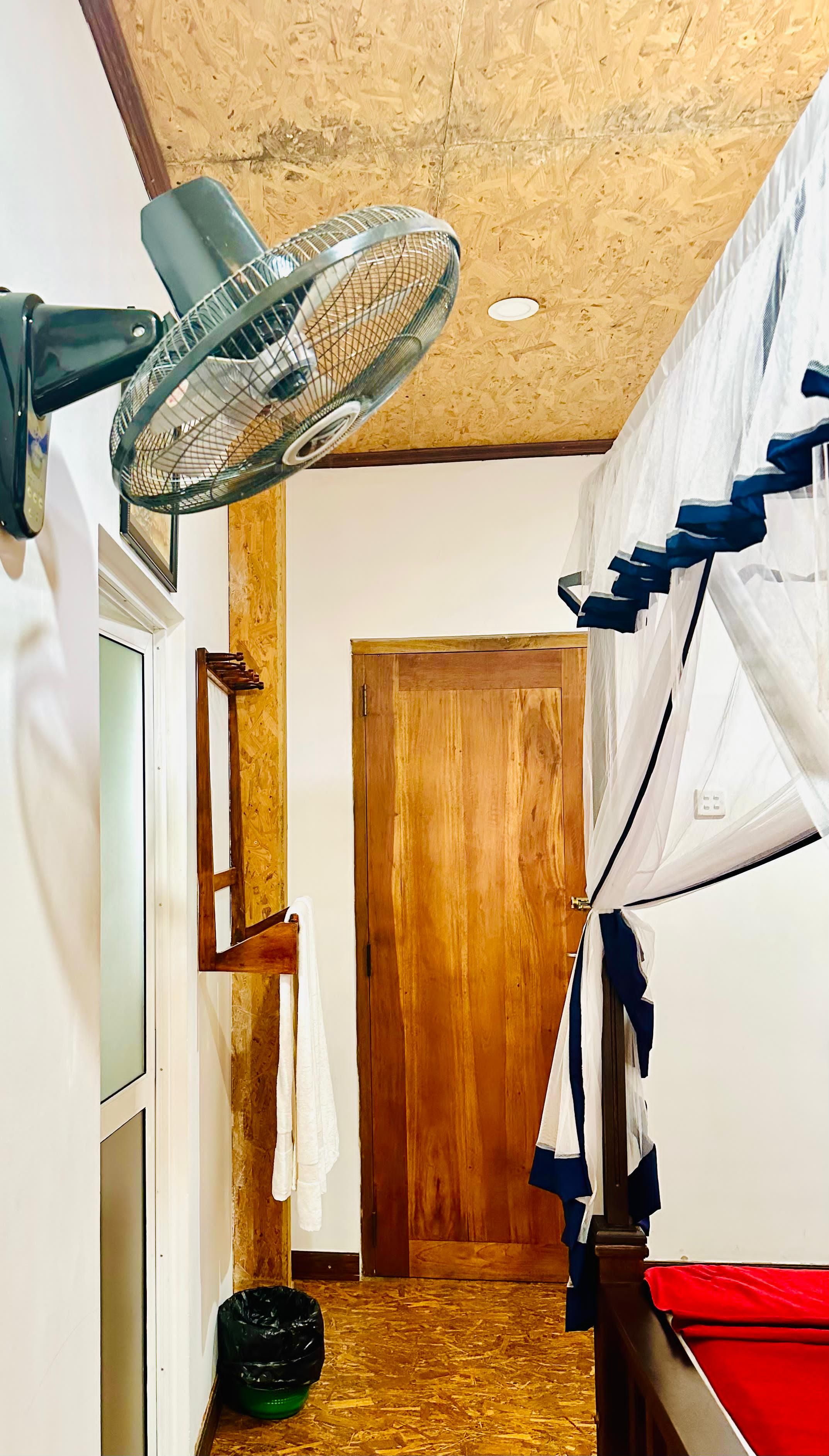 A room with a wooden door, a fan, a bed with a white and blue canopy, and a small black trash bin.