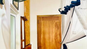 A room with a wooden door, a fan, a bed with a white and blue canopy, and a small black trash bin.