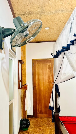 Ein Zimmer mit Holztür, einem Ventilator, einem Bett mit weißblauer Baldachin und einer kleinen schwarzen Mülltonne.