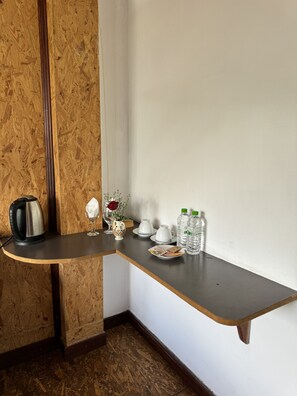 Een hoek van een kamer met een houten vloer, een donkerbruine tafel met een waterkoker, een vaas met bloemen, een glas en twee waterflessen.