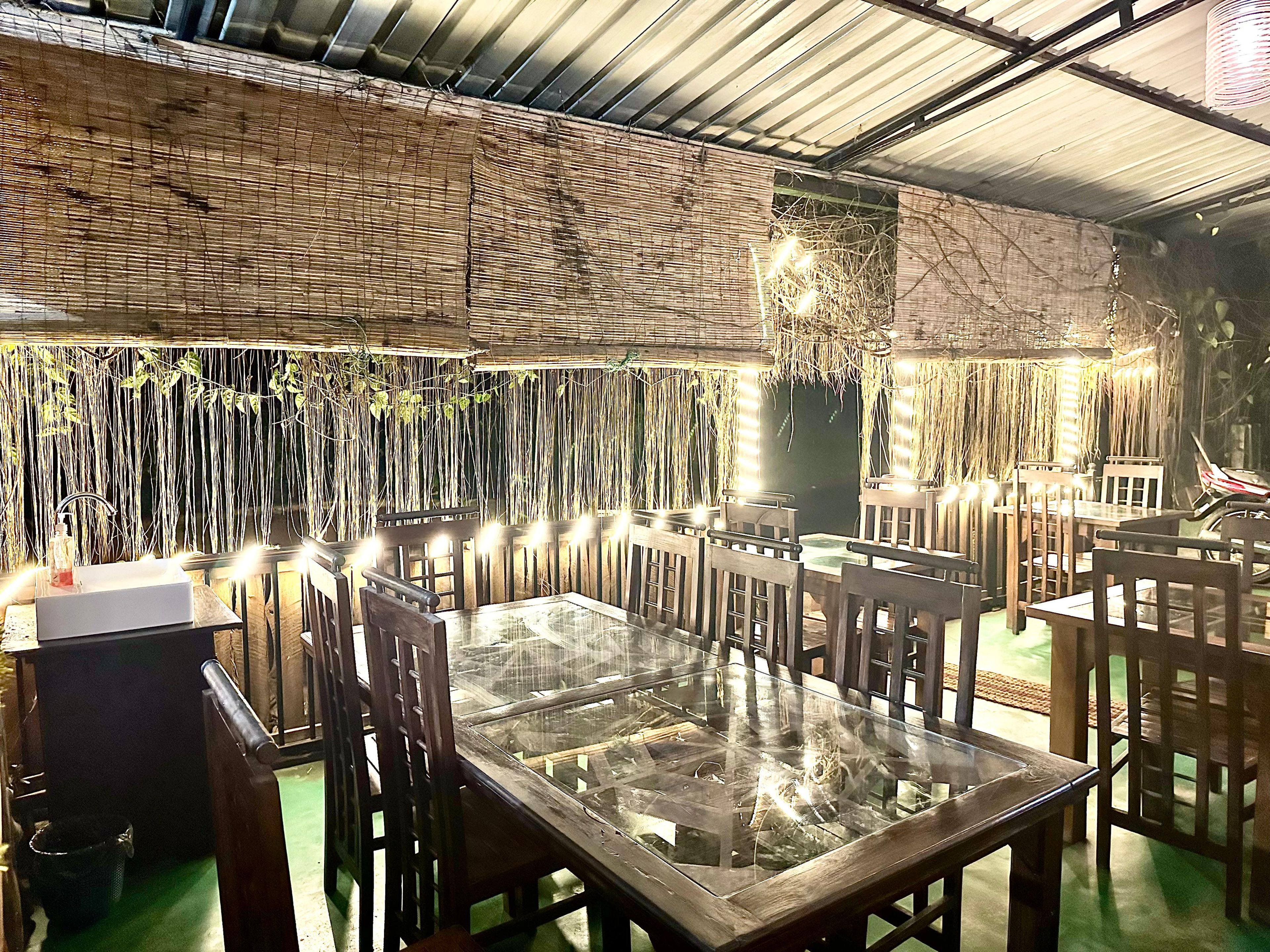 Un espace de restauration intérieur avec des tables et des chaises en bois, un mur recouvert de chaume et des guirlandes lumineuses.