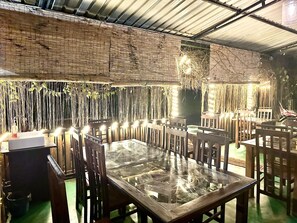 An indoor dining area with wooden tables and chairs, a thatched wall, and string lights.