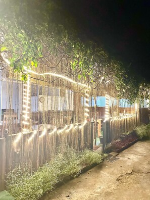 A garden area with a fence adorned with string lights, a wooden bench, and a metal grate on the ground.