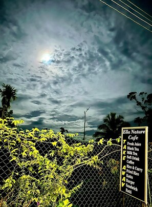 Un cartel que enumera los artículos del menú de Ella Nature Cafe, con un cielo nublado de fondo y una cerca con follaje verde.