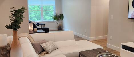 A modern living room with a large sectional sofa, a wooden coffee table, and a dining table. There is a large window providing natural light and a view of greenery outside.