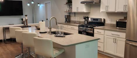 A modern kitchen with a central island, bar stools, stainless steel appliances, and a ceiling light fixture.