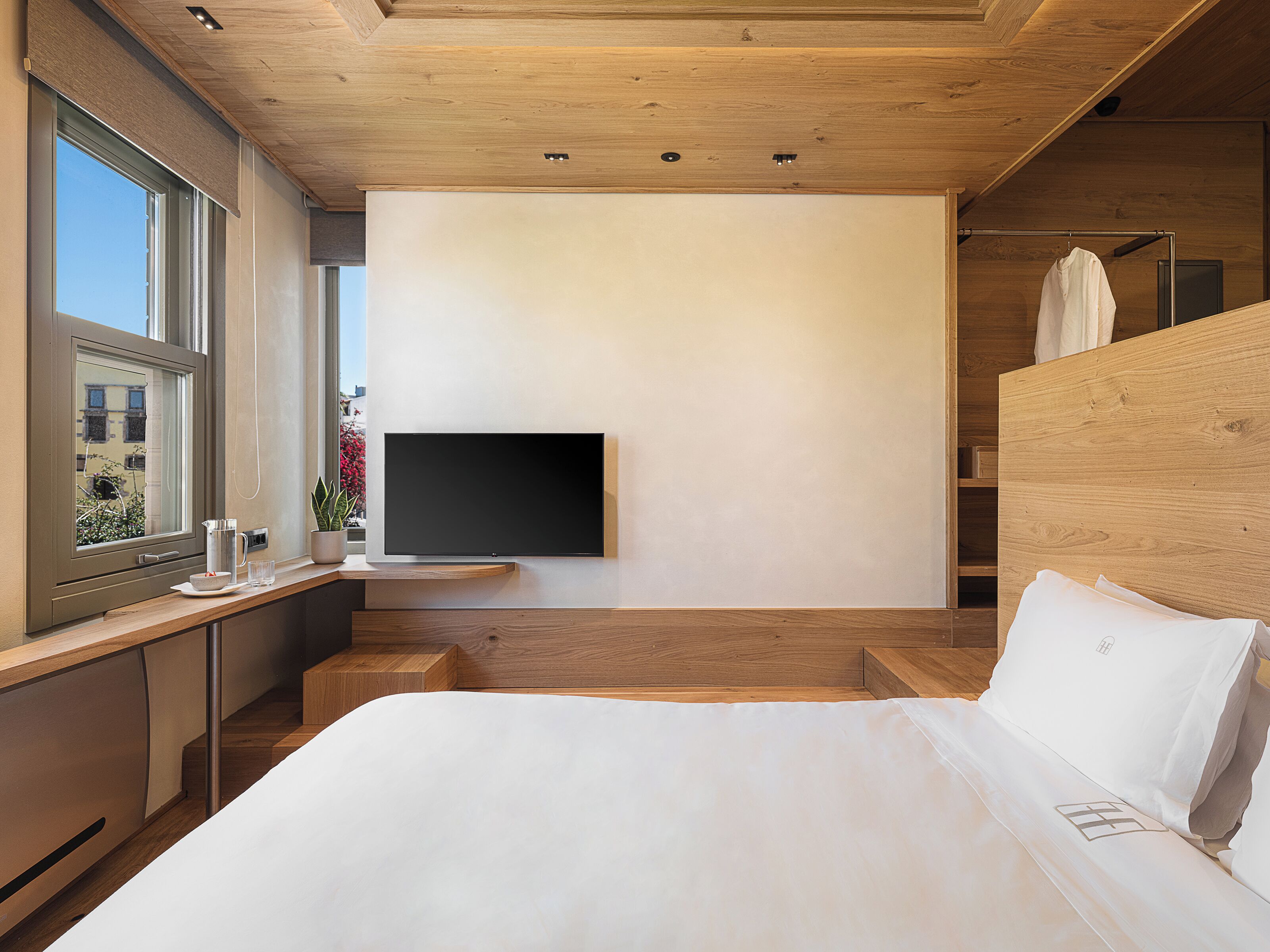 Un dormitorio moderno con una cama grande, un televisor montado y una ventana con vistas a edificios y flores.