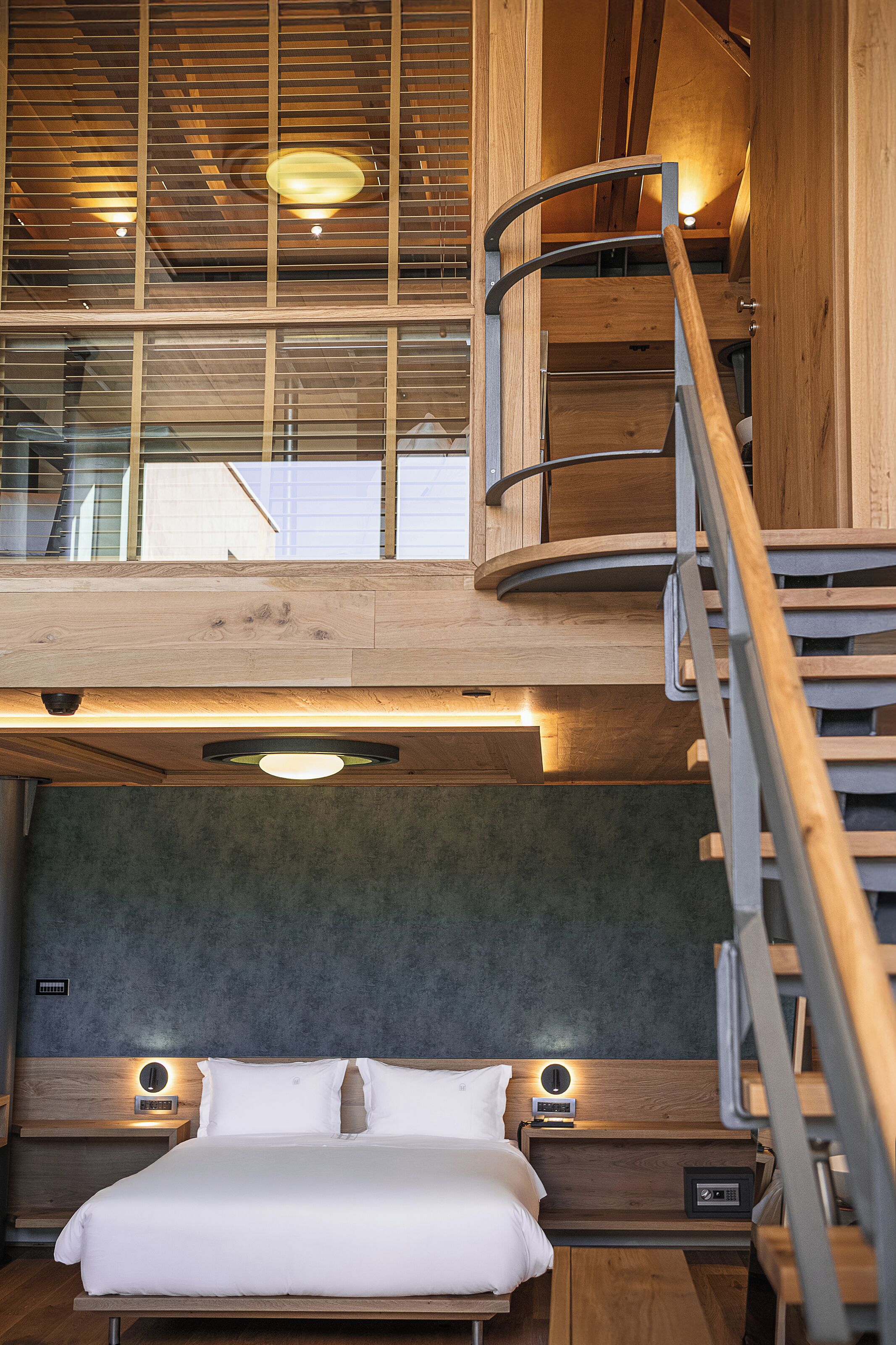 A modern hotel room with a lofted sleeping area, a wooden staircase, and a bed with a headboard.