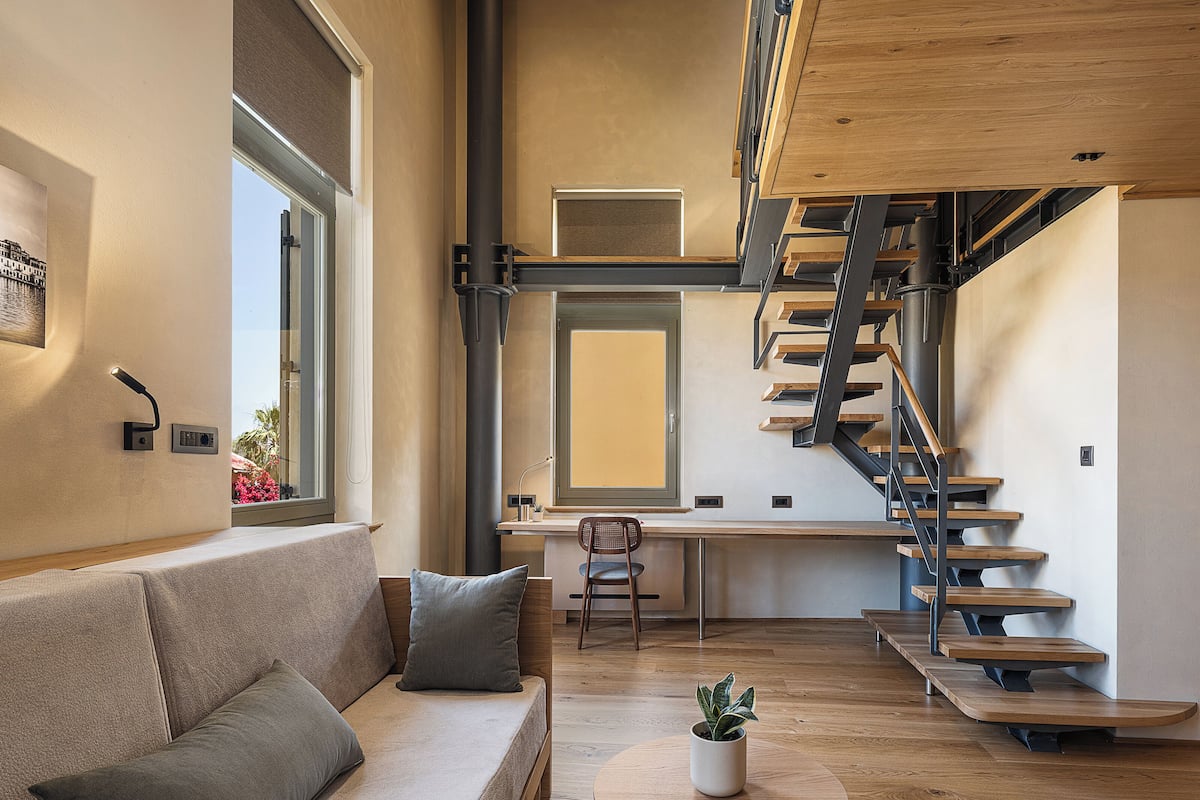 Een modern interieur met een houten trap, een bank met kussens, een kleine tafel met een plant en een raam met jaloezieën.