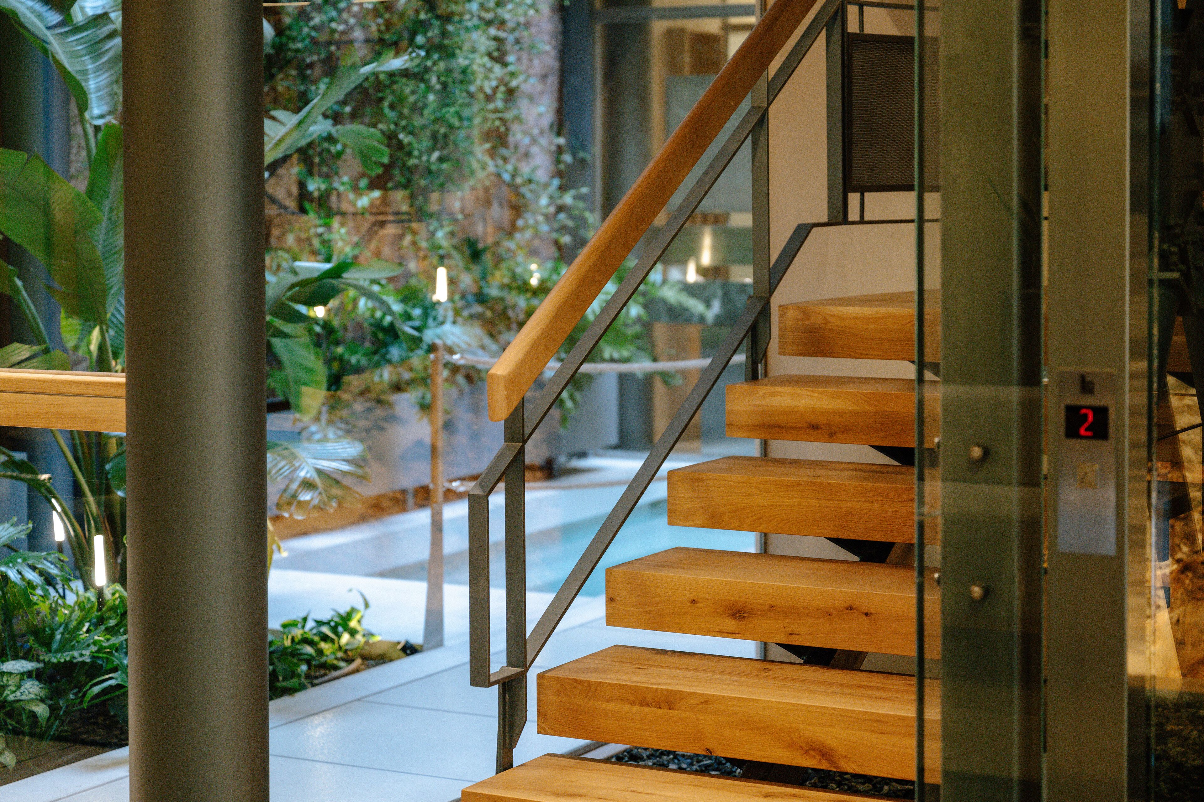 A modern staircase with wooden steps and metal railings, leading to an indoor area with plants and a glass wall.