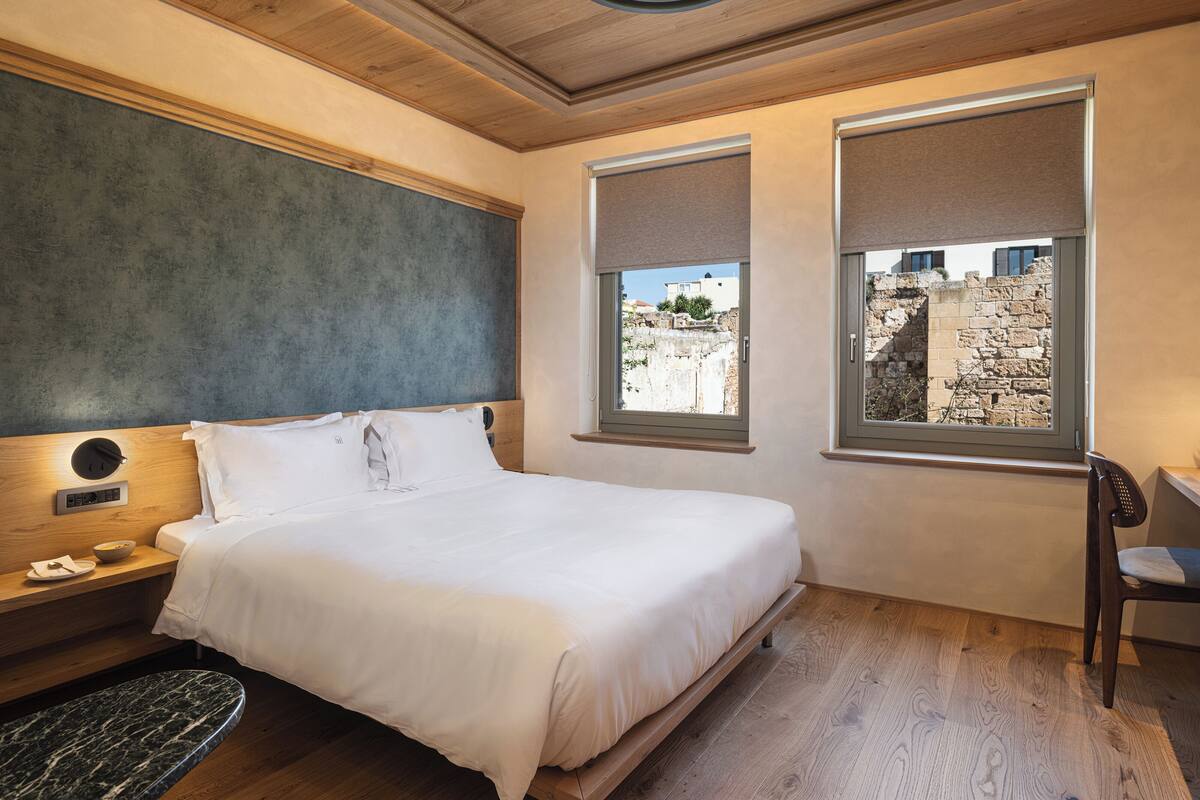 Een moderne slaapkamer met een houten plafond, een bed met witte lakens, een nachtkastje en uitzicht op een stenen muur door een raam.