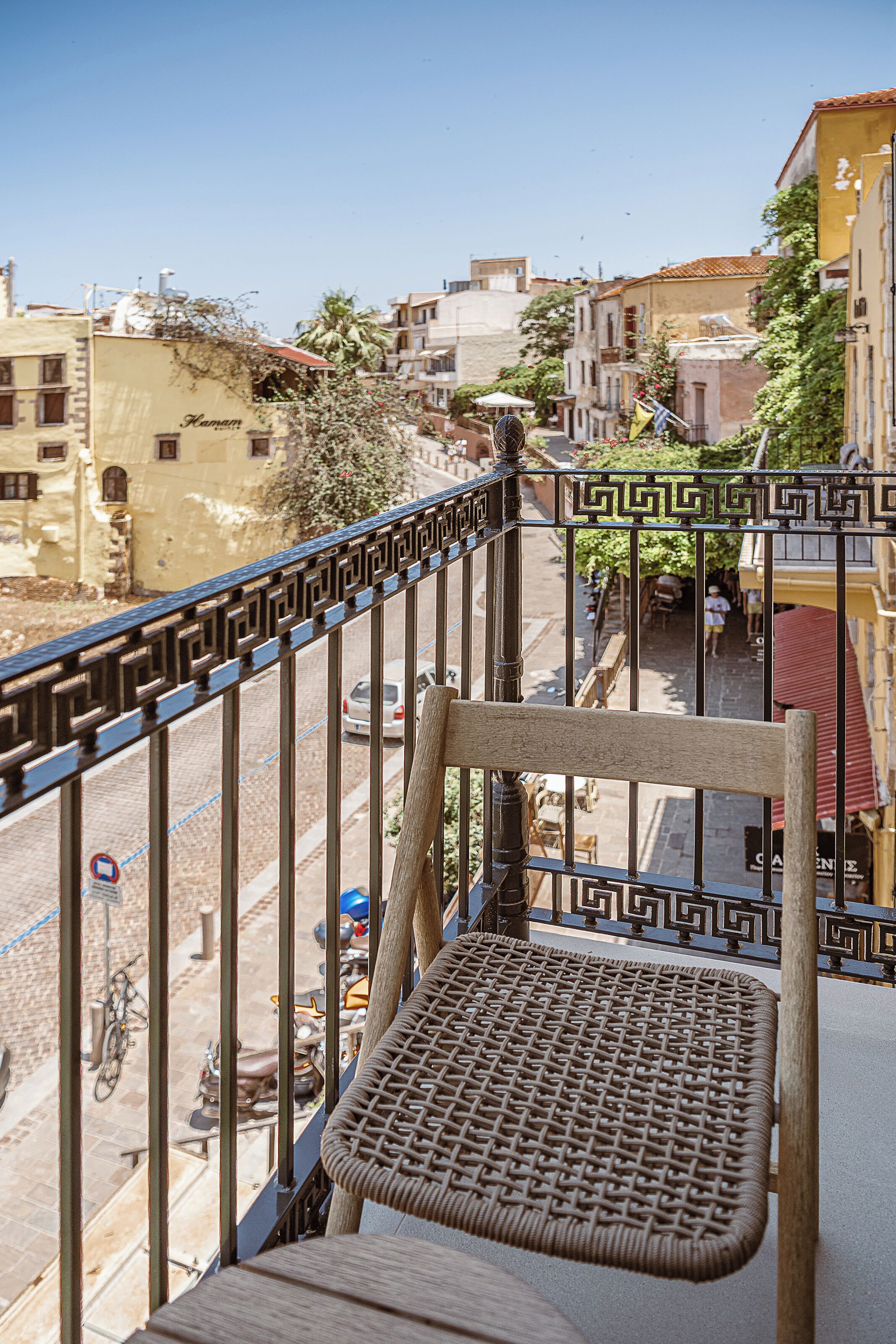 Un balcone con una sedia in vimini che si affaccia su una strada con edifici e veicoli parcheggiati.