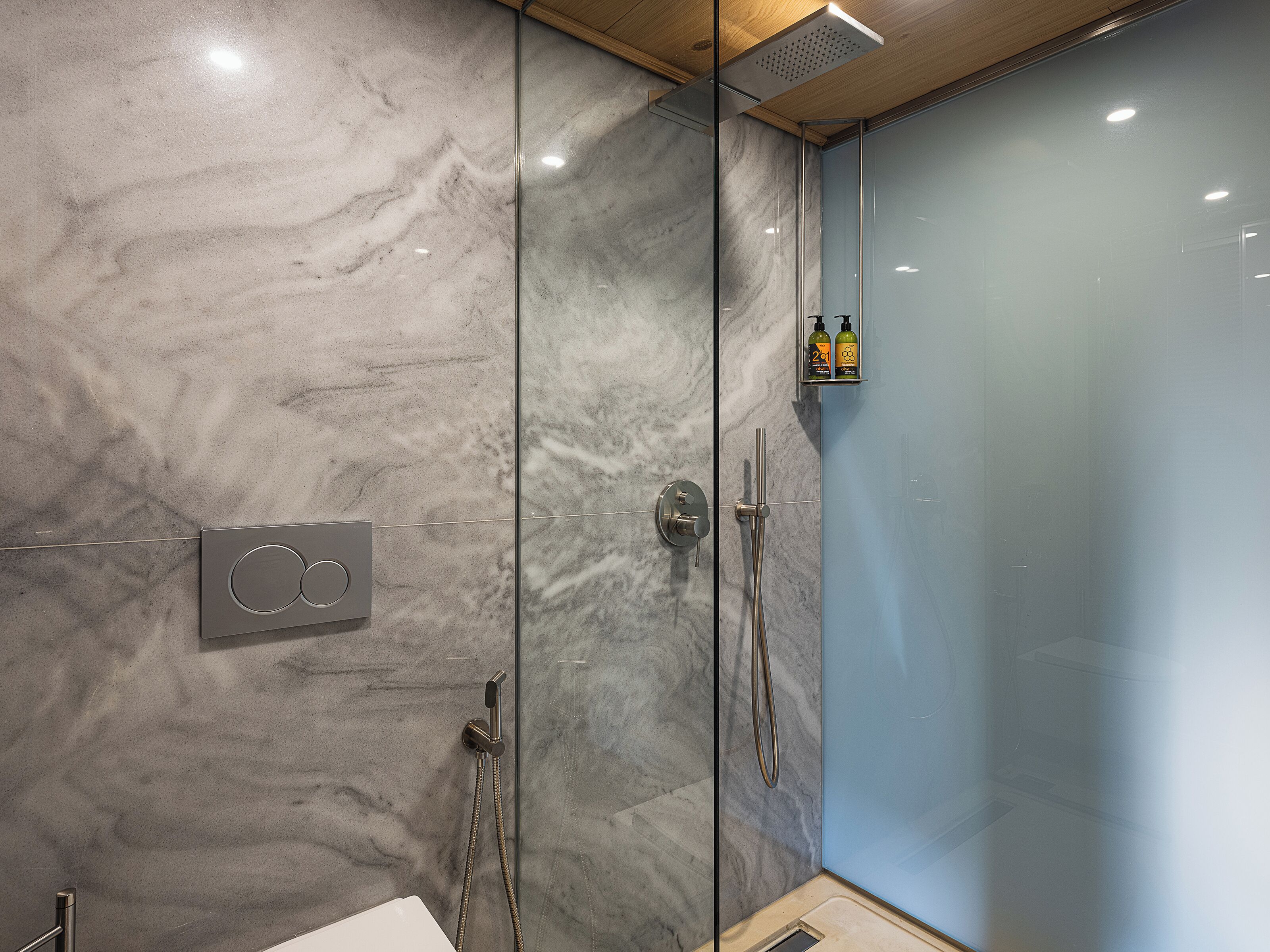 A modern bathroom with a glass shower enclosure, a marble wall, and a small shelf with toiletries.