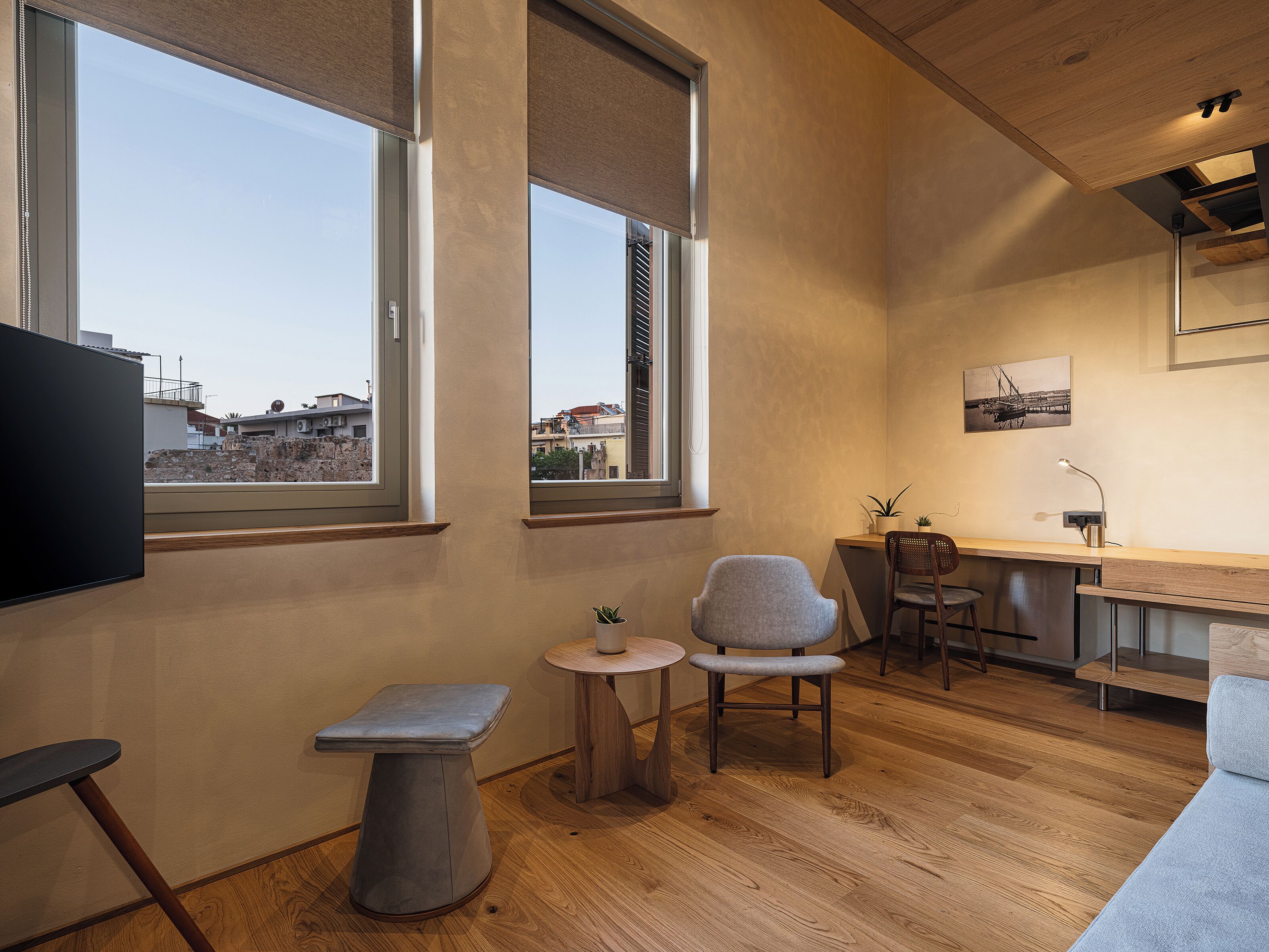 Une pièce moderne et bien éclairée, dotée de grandes fenêtres, d’une petite table ronde, de deux chaises, d’un banc et d’un bureau avec une lampe.