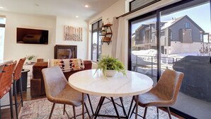 Un coin repas moderne avec une table ronde, deux chaises, une plante en pot et une grande fenêtre donnant sur une maison et un paysage enneigé.