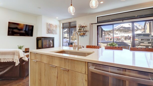 Une cuisine moderne dotée d’un grand îlot central, de meubles en bois, d’un lave-vaisselle en acier inoxydable, de luminaires suspendus et offrant une vue sur une montagne enneigée.