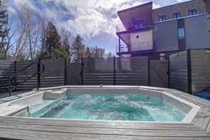 Uma banheira de hidromassagem com deck de madeira, cercada por uma grade de metal, e um edifício moderno com varandas ao fundo.