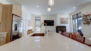 Une cuisine moderne dotée d’un grand îlot central, d’appareils électroménagers en acier inoxydable, de luminaires suspendus et d’un coin salon confortable avec une cheminée.